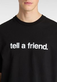 Sort bomulds t-shirt med rund halsudskæring prydet med teksten "fortæl en ven" i fed hvide bogstaver på tværs af brystet.