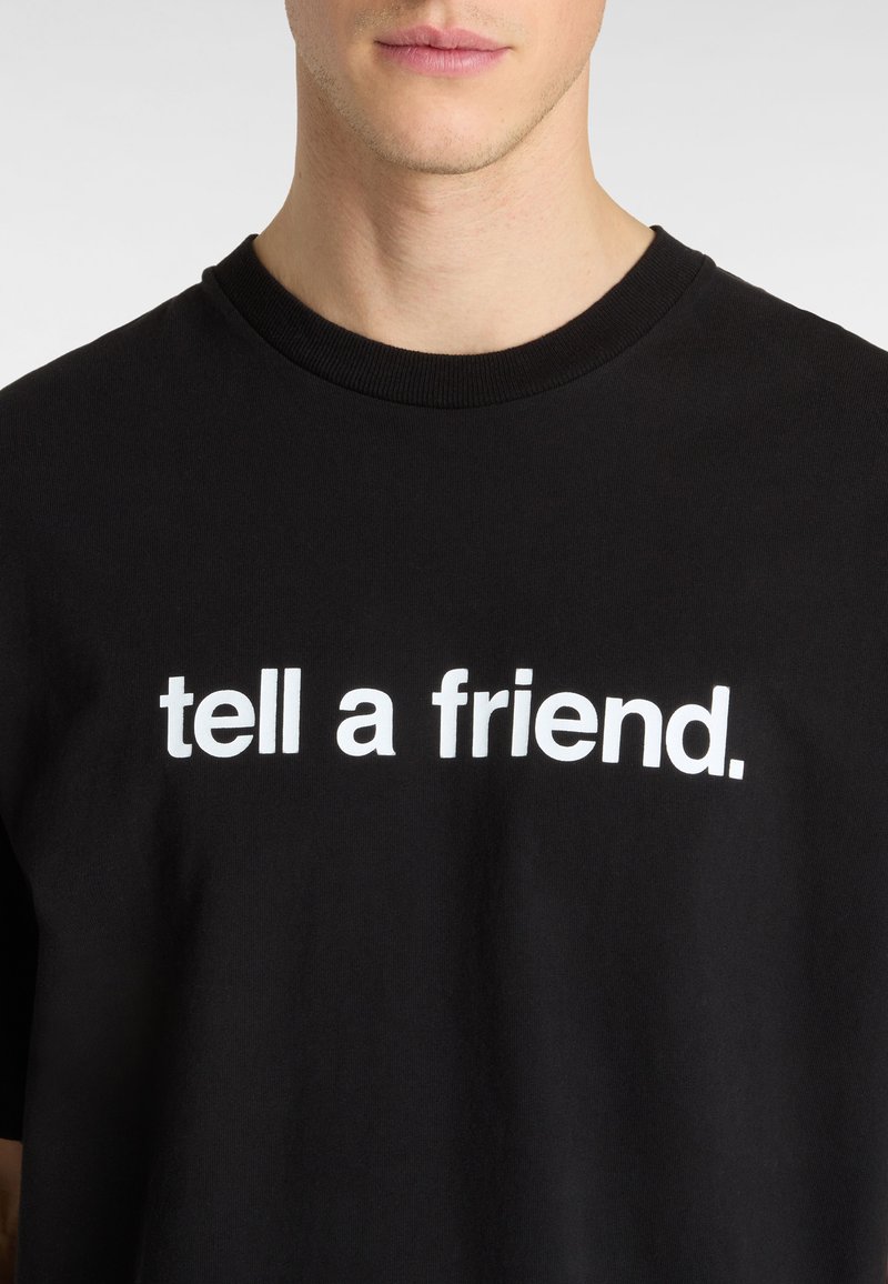 T-shirt en coton noir avec un col rond portant le texte "dis à un ami" en lettres blanches en gras sur la poitrine.