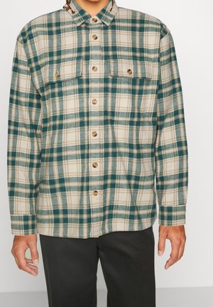 Man draagt een groen-beige geruit overhemd met klepzakken op de borst, gecombineerd met een zwarte broek, tegen een effen achtergrond.