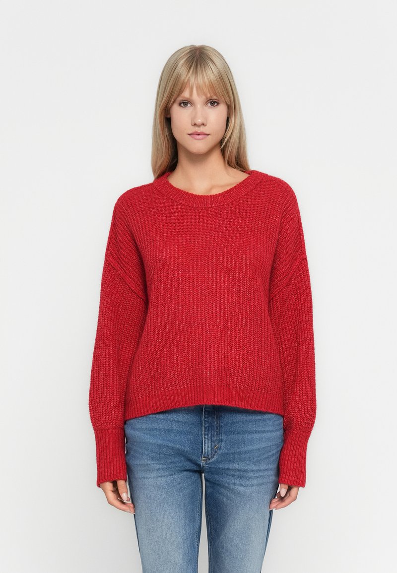 Rød strikkegenser med vid halslinning og senkede skuldre. Den har en løs passform og ribbestrikkede mansjetter, kombinert med blå jeans.