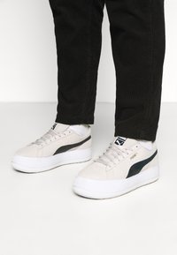 Baskets en suede blanches avec une bande d'accent noire, semelle en caoutchouc épaisse et design à lacets, associées à un pantalon noir côtelé.