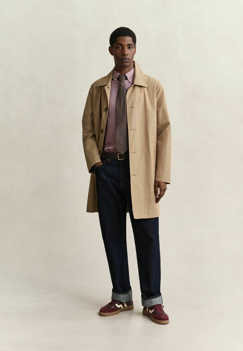 Jeune homme portant un trench-coat beige, une cravate à motifs, une chemise rose, un jean foncé avec revers et des baskets rouges, debout devant un fond uni.