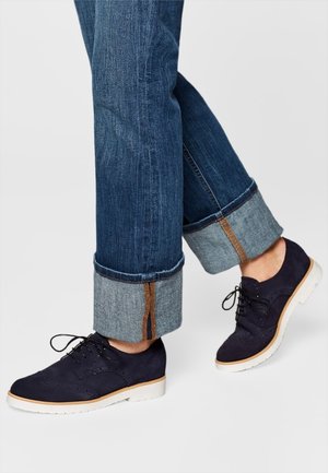 Personne portant un jean bleu en denim retroussé et des chaussures à lacets bleu marine foncé avec des semelles blanches, debout sur une surface claire.