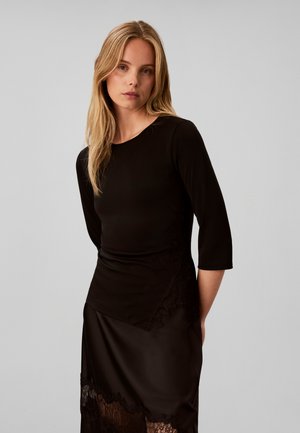 Jeune femme aux longs cheveux blonds portant un haut noir à manches longues et une jupe noire avec des détails en dentelle, debout devant un fond gris uni.
