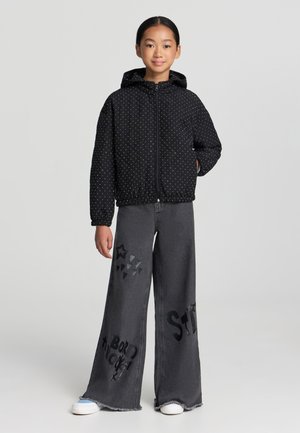 Giacca nera con zip, decorata con pois bianchi e cappuccio, abbinata a pantaloni in denim grigio larghi con motivi stampati e orli sfilacciati.