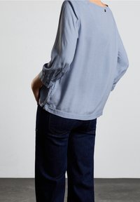 Ljusblå långärmad topp med rund halsringning och detalj med knapp i ryggen, kombinerad med mörk denimjeans med avslappnad passform.