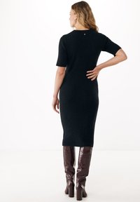 Černé žebrované pletené šaty s krátkými rukávy a přiléhavým střihem, doplněné lesklými hnědými kozačkami po kolena. Zadní pohled odhaluje jemný detail výstřihu.