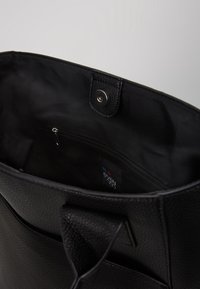 Mustan oljetasku, jossa on teksturoitu pinta. Sisäpuolella on vetoketjullinen tasku ja logotagi. Laukku on muotoiltu ja siinä on tukevat kahvat.