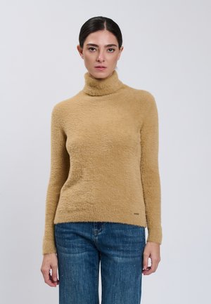 Donna con capelli scuri raccolti indossa un maglione a collo alto beige e peloso e jeans blu su uno sfondo grigio semplice.