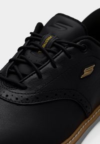 Fekete bőr fűzős cipő arany "S" logóval, perforált részletekkel és barna talpéllel, közeli felvétel az elülső és oldalsó részekről.