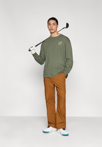 Μπλούζα μακριάς μανίκας ελιάς με λογότυπο, καφέ παντελόνι και λευκά παπούτσια γκολφ με μπλε λεπτομέρειες, κρατώντας ένα γκολφ κλαμπ στον ώμο.