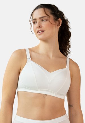 Woman with dark curly hair wearing a white soft-cup bra with adjustable straps, shown from waist up against a plain background.