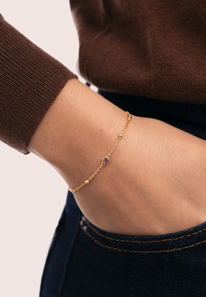 Pulsera de cadena de oro con pequeños detalles esféricos y una gema central de amatista. Lleva puesta en una muñeca, mostrando piel y un suéter marrón.