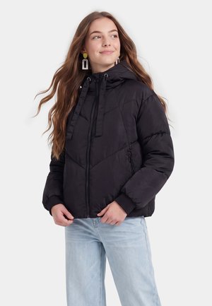 Giovane donna con capelli lunghi che indossa una giacca imbottita nera con cappuccio e jeans azzurri chiari, guarda di lato e sorride leggermente.