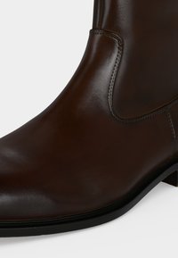 Gros plan sur une bottine en cuir marron foncé avec des coutures visibles et une semelle noire sur un fond clair uni.