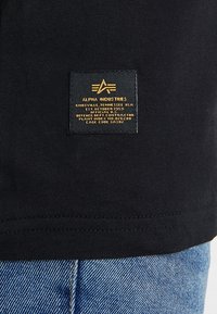 Černé bavlněné tričko s vyztuženým lemem a žlutou etiketou s názvem značky "Alpha Industries" a různými výrobními detaily.