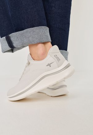 Primer plano de una persona que lleva zapatillas gris claro con una franja y un logo plateado, combinadas con unos jeans oscuros de puños sobre un suelo blanco.