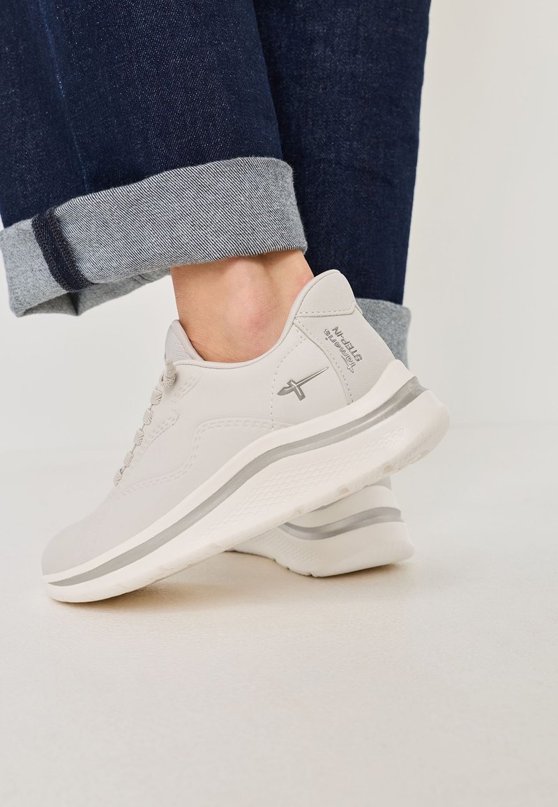 Ingrandimento di una persona che indossa sneakers grigio chiaro con una striscia e un logo argentati, abbinate a jeans blu scuro con risvolto su un pavimento bianco.