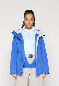 Μπλε μόνωτο μπουφάν με γκρι κουκούλα, που διαθέτει πολλαπλές τσέπες και αντίθετη ραφή. Φορεμένο πάνω από ένα ανοιχτό μπλε τοπ και μπεζ παντελόνι.