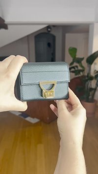 Juodos odos rankinė su struktūruota forma, priekyje turinti auksinę metalinę sagtį ir logotipo akcentą. Tekstūruota paviršius su vertikaliomis siūlėmis.