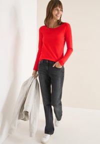 Rotes Langarmoberteil mit Rundhalsausschnitt, kombiniert mit dunkelgrauen Jeans. Die Person hält einen grauen Pullover und trägt weiße Sneaker.