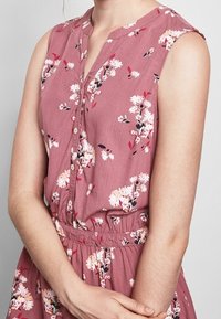 Blommig klänning i dammros med knappstängning fram, ärmlös design, elastisk midja och blommönster i vitt och rosa.