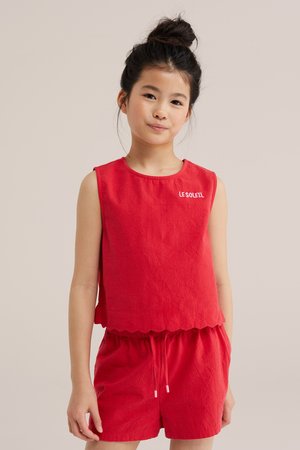 Niña joven con el pelo oscuro recogido en un moño, que lleva una camiseta roja sin mangas con el texto "LE SOLEIL" y pantalones cortos rojos a juego sobre un fondo claro.