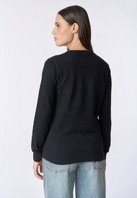 Maglione nero a maniche lunghe e collo tondo, realizzato in un tessuto texturizzato. La parte posteriore è semplice con un orlo leggermente più lungo. Abbinato a jeans azzurri chiari.