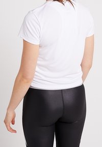 Vit kortärmad träningsskjorta i kombination med svarta glänsande leggings. Skjortan har en rundad halsringning och leggingsen har en slät och smidig textur.
