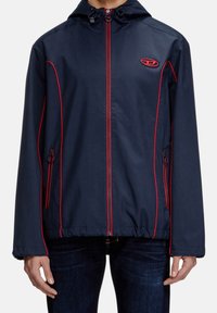 Granatowa kurtka z czerwonymi lamówkami, z pełnym zamkiem, regulowanym kapturem, bocznymi kieszeniami i logo na klatce piersiowej. Gładki, lekki materiał.