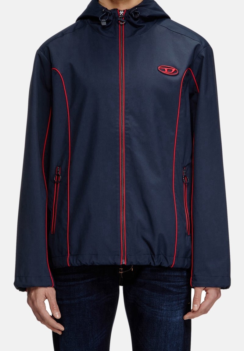 Granatowa kurtka z czerwonymi lamówkami, z pełnym zamkiem, regulowanym kapturem, bocznymi kieszeniami i logo na klatce piersiowej. Gładki, lekki materiał.