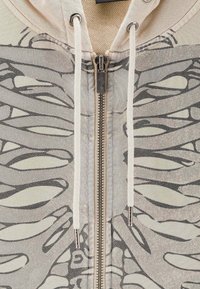 Felpa con zip in tessuto beige con un motivo astratto grigio. Presenta cordini e accessori in argento. Texture morbida.