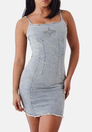 Femme portant une robe en denim bleu clair cintrée avec de fines bretelles, un détail en forme d'étoile sur la poitrine et un ourlet effiloché, debout devant un fond uni.
