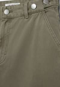 Detaljni prikaz maslinasto zelenih traperica sa srebrnim gumbima i vidljivim šavovima na prednjim džepovima, prikazuje teksturu tkanine i etiketu na pojasu.