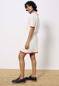 Weißes Strickkleid mit rotem Saum am Ausschnitt und am Saum, mit einem taillierten Oberteil und einem ausgestellt Rock. Getragen mit dunklen Schuhen und sichtbarem Tattoo.