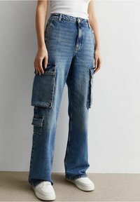 Pantalones de mezclilla cargo de pierna ancha con múltiples bolsillos de parche, lavado azul claro, dobladillos deshilachados y un ajuste relajado. Combinados con zapatillas blancas.
