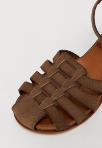 Sandal en suède marron avec des sangles tressées, un bout rond et une semelle plate. L'intérieur est en cuir beige clair contrastant.