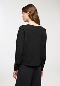 Kobieta z długimi brązowymi włosami stoi tyłem, ubrana w luźny czarny sweter i czarne spodnie na białym tle.