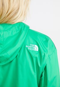 Zelená bunda s kapucí, vyrobená z lesklého materiálu. Na levém rameni se nachází bílé logo "The North Face." Hladký povrch.