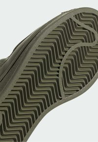 Detailní záběr na olivově zelenou podešev boty se zigzagovým vzorem a texturovaným gumovým okrajem.