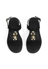 Schwarze Wildledersandalen mit flacher Sohle, die zwei reflektierende goldene runde Akzente und einen verstellbaren Knöchelriemen besitzen.