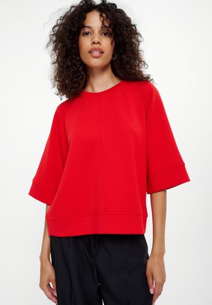 Red, short-sleeved top made of soft material with a relaxed fit, featuring a round neckline and visible seam detailing, paired with black pants.