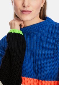 Strickpullover mit blauem Körper, schwarzen Ärmeln und leuchtend grünen Bündchen. Verfügt über eine gerippte Textur und ein Design mit Farbsperren, einschließlich Orange.