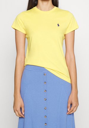 Mujer que lleva una camiseta amarilla ajustada de manga corta con un pequeño logo azul, metida dentro de una falda azul acanalada de talle alto con botones en el frente.