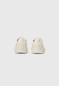 Para off-white butów sportowych widziana z tyłu, z teksturowanymi podeszwami i uchwytami na pięcie, na jednolitym jasnym tle.