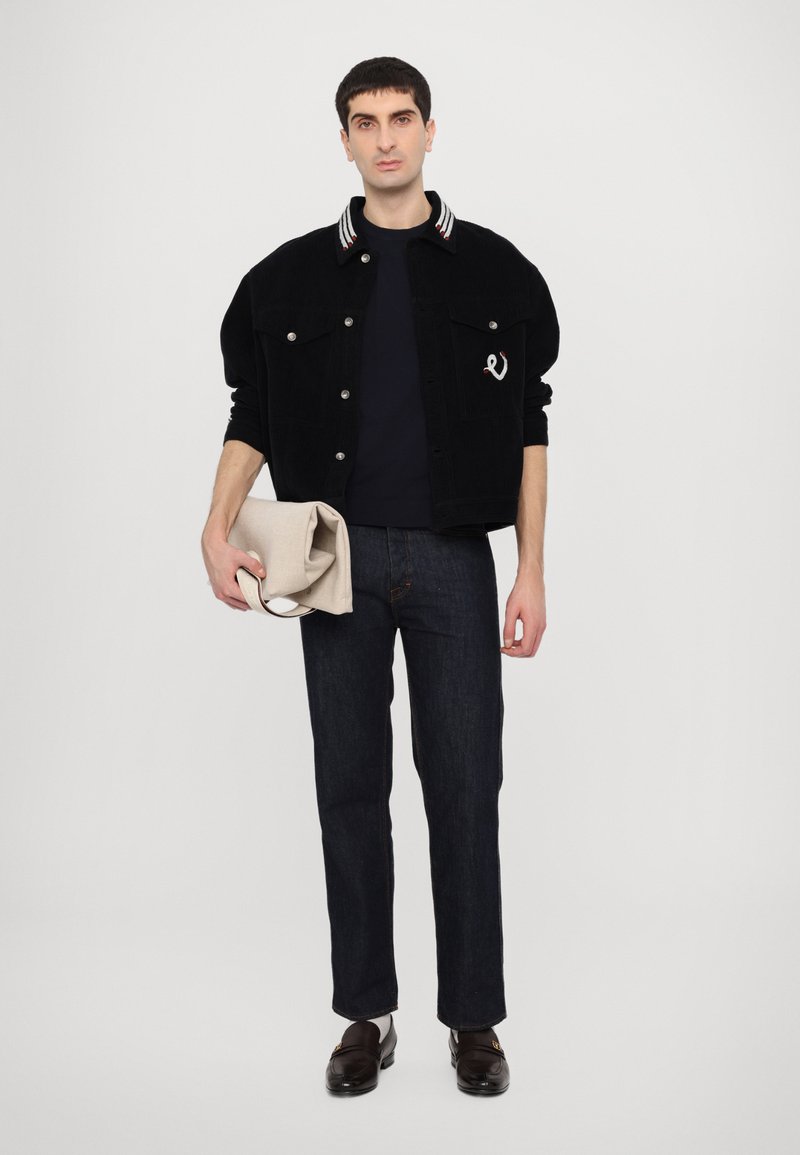 Giacca in velluto a coste nera con colletto a righe, t-shirt scura, jeans di denim scuro e borsa beige piegata. Il modello indossa scarpe marroni.