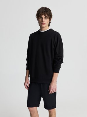 Giovane uomo con capelli castani ondulati che indossa una felpa nera a maniche lunghe sopra una camicia bianca e pantaloncini neri fino al ginocchio, in posizione neutra.