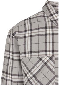 Camicia a quadri grigia, bianca e nera nella zona delle spalle e del petto con colletto piegato e taschino su uno sfondo neutro.