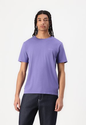 T-shirt en coton violet à manches courtes, col ras du cou, avec un petit logo sur la poitrine, associé à un jean bleu foncé.