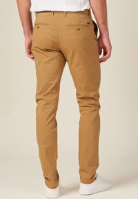 Beige Chinos in schlanker Passform, zwei Gesäßtaschen, minimalistische Nähte und kombiniert mit weißen Sneakers. Glattes Baumwollmaterial.